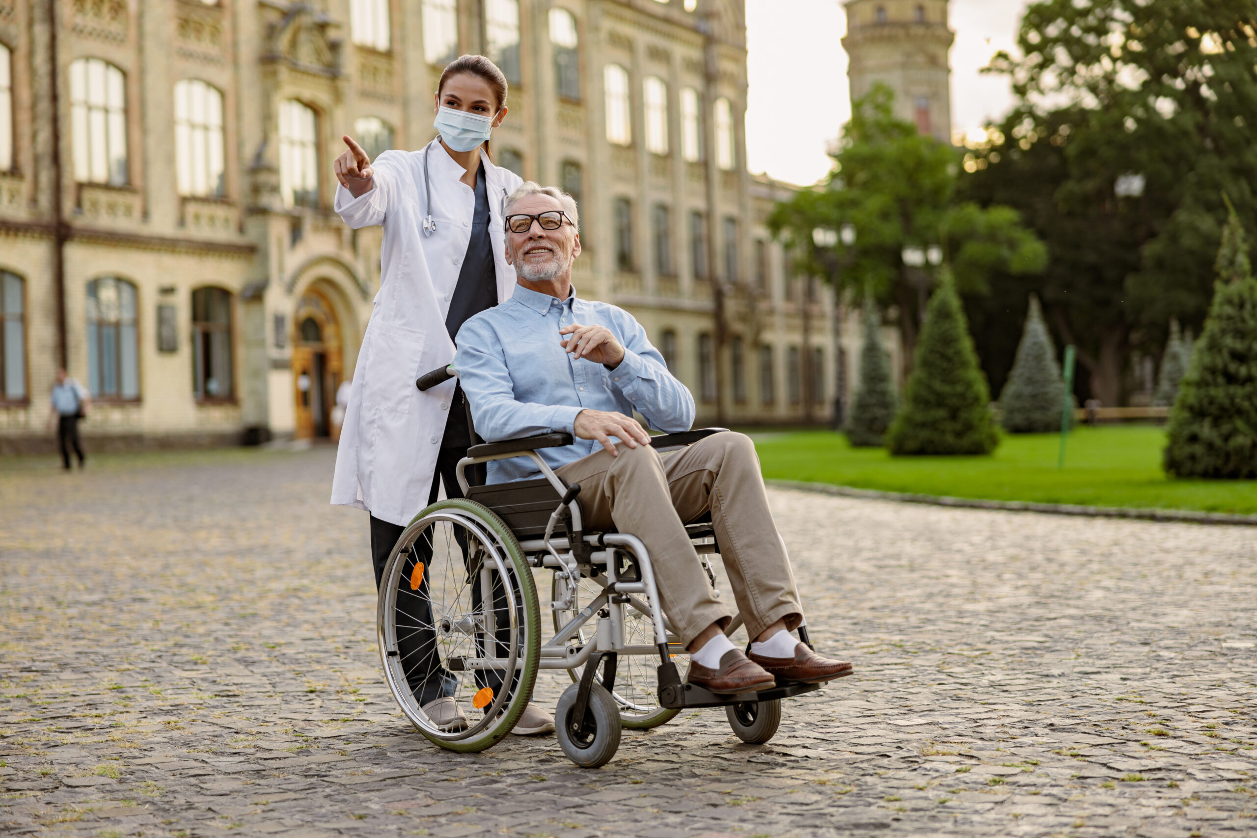 Full length shot of caring young nurse in protective mask taking care of senior man handicapped patient in wheelchair outdoors. Health care, occupation concept